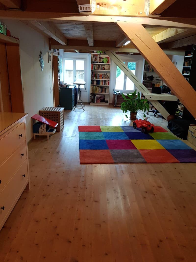 Bright, open room with wooden floors, exposed wooden beams, large windows, bookshelf, desk, chair, plant, toy car, colorful rug, and small wooden stool.