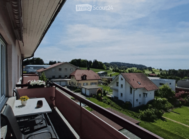 Balcony with a table, chairs, and flower pots, overlooking a scenic countryside with houses and green fields.