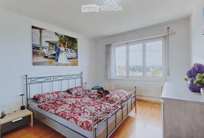 Bright bedroom with wooden flooring, large windows, a metal-framed bed with a red and white patterned duvet, a nightstand with a lamp, and a large picture frame on the wall.