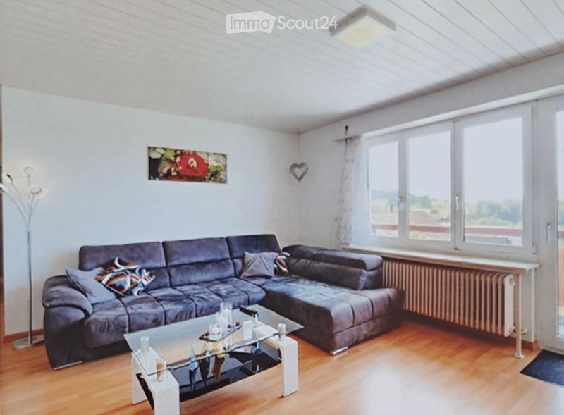 Modern living room with a grey sectional sofa, glass coffee table, floor lamp, and a picture frame on the wall. The room has wooden flooring and large windows with a view of the outside.