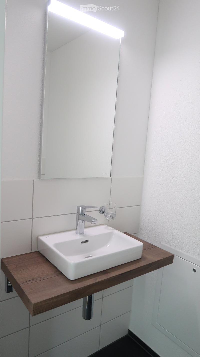 Modern bathroom with a white sink on a dark wooden countertop, a chrome faucet, a large mirror with built-in lighting, and a glass cup on the countertop.