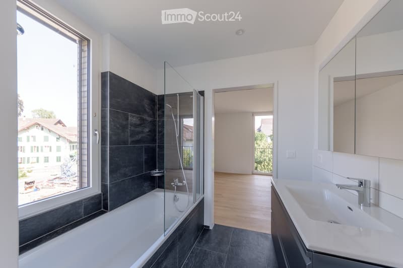 Modern bathroom with a bathtub, glass shower enclosure, sink, and mirror. Large window offers a view of the surrounding area.