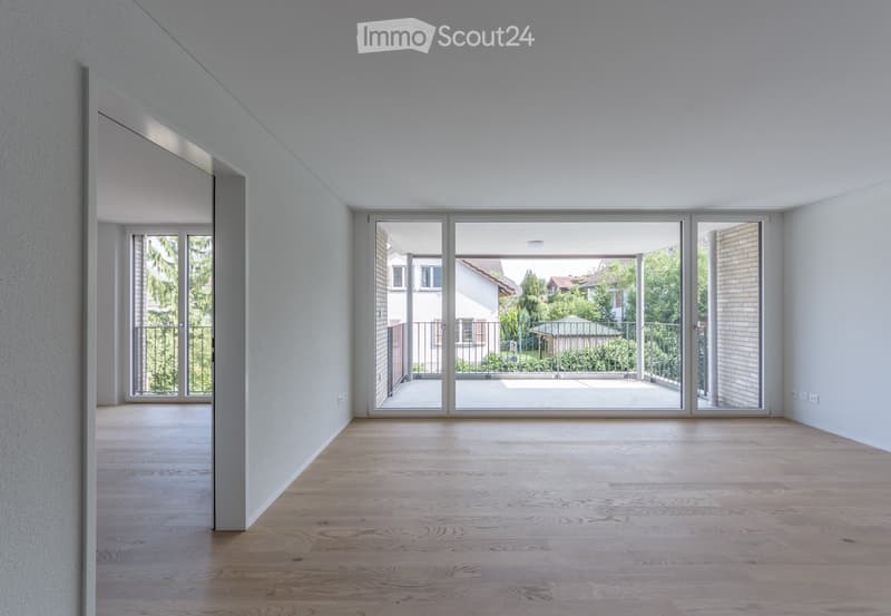 Empty room with light wooden flooring, white walls, large sliding glass doors leading to a balcony, and a view of greenery outside.