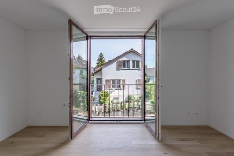 Empty room with wooden flooring and large glass doors leading to a balcony or terrace, overlooking a residential area with houses and greenery.