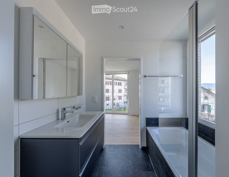Modern bathroom with a sink, faucet, large mirror, and bathtub, featuring a view of the balcony and cityscape through large windows.