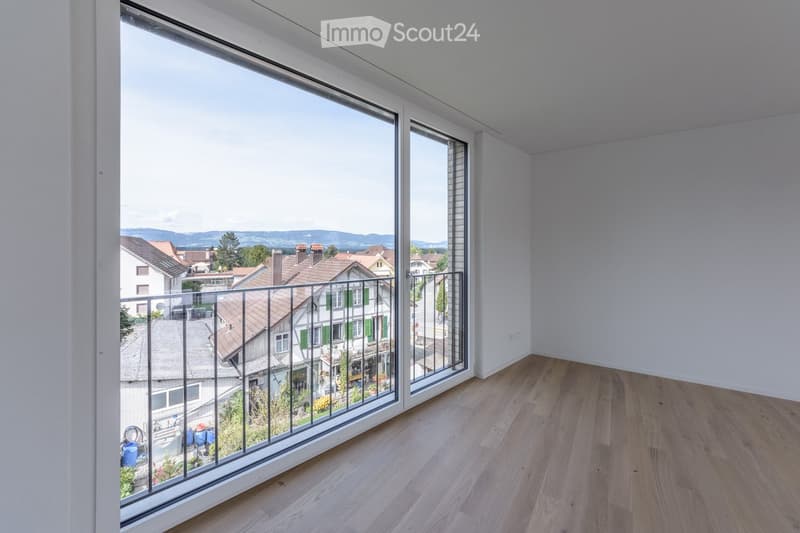 Empty room with wooden flooring, large windows, and a metal railing leading to a view of a town and surrounding landscape.