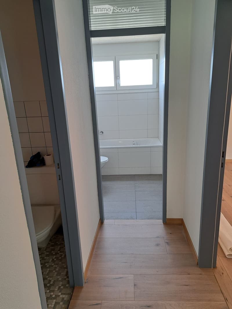 A narrow hallway with wooden flooring leads to a bathroom. The bathroom has white tiled walls, a bathtub, and a toilet. The hallway has light-colored walls and a small window at the end.