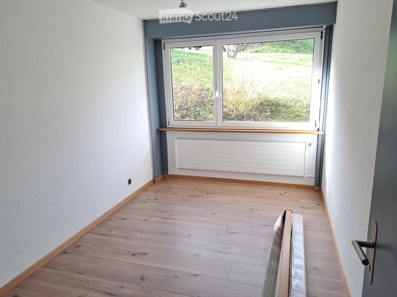 Empty room with light wooden flooring, white walls, a radiator, and a large window with a view of a green outdoor area.