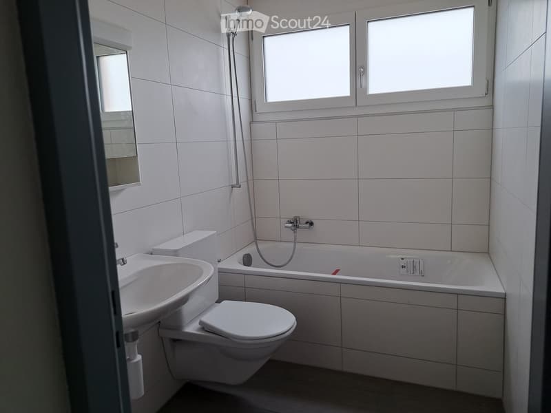 Modern bathroom with white tiles, bathtub, toilet, sink, and mirror. Features a showerhead and window for natural light.