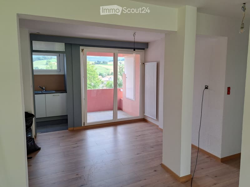 Empty room with wooden flooring, white walls, and a sliding glass door leading to a balcony with a view of a green landscape.