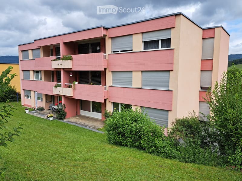 A modern two-story building with pink and beige exterior, multiple windows, balconies, and surrounding greenery.