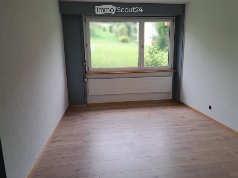 Empty hallway with wooden flooring, white walls, a window with a view of greenery, and a radiator below the window.