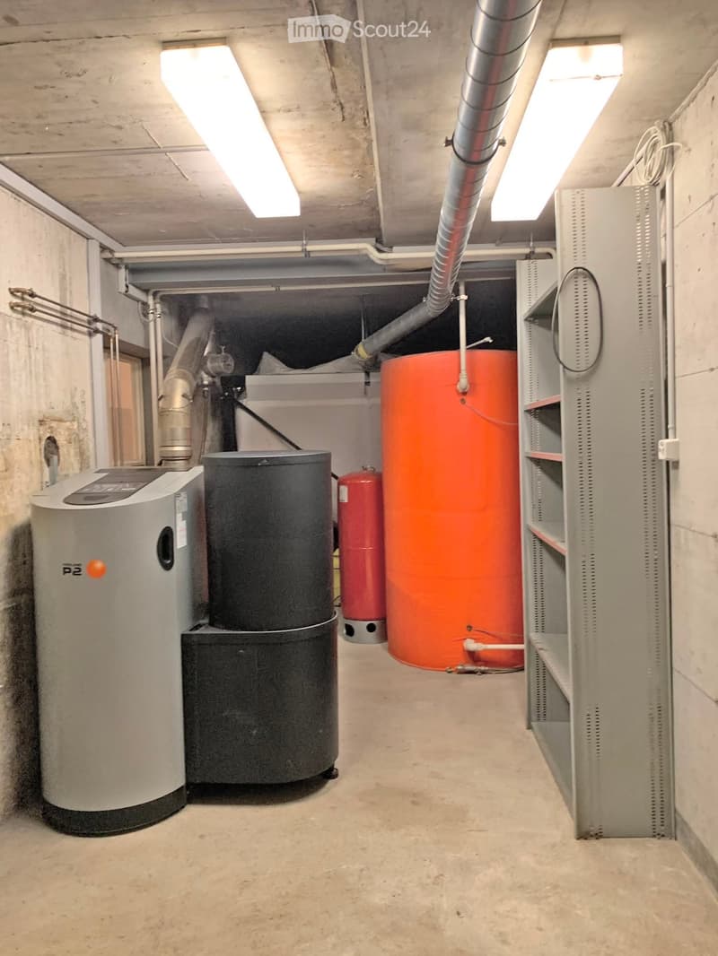 Utility room with water tank, boiler, and electrical panel, concrete walls and floor, fluorescent lighting.