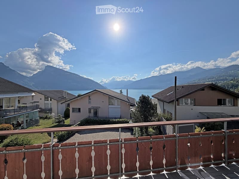 Balcony overlooking a scenic view of mountains, a lake, and neighboring houses. The balcony features a wooden railing and is situated on a raised ground floor.