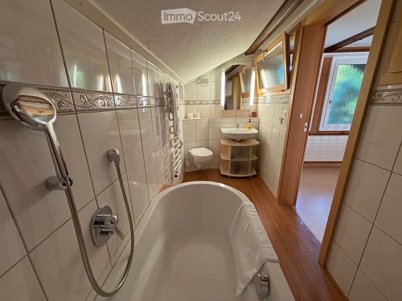 Modern bathroom with a bathtub, toilet, sink, mirror, and a showerhead. The room features tiled walls and wooden flooring.