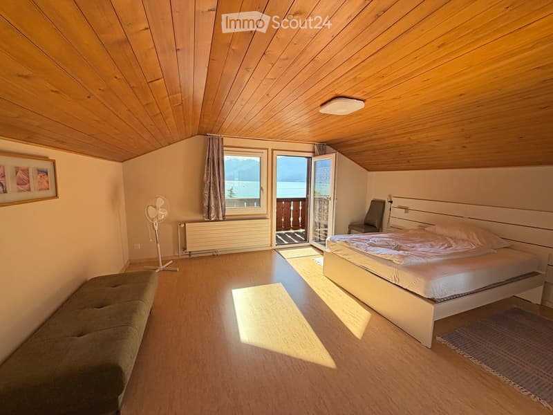 Cozy attic bedroom with wooden slanted ceilings, large window, and open sliding door leading to a balcony. Features a bed, sofa, fan, chair, radiator, and picture frame on the wall.