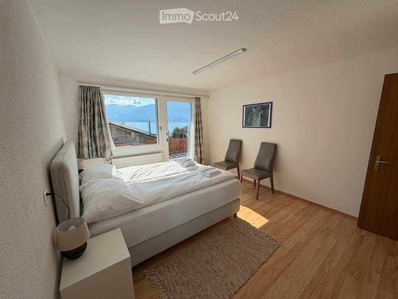 Cozy bedroom with a double bed, nightstand, lamp, two chairs, and a scenic mountain view through large windows.