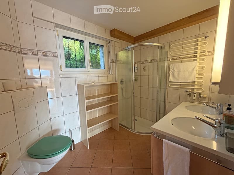 Modern bathroom with a green toilet seat, double sink, shower, and built-in wooden shelf. Features tiled walls and a window with a view of greenery.