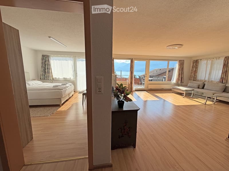 Bright, open-plan living and bedroom area with wooden flooring, large windows offering a scenic view, a sofa, coffee table, bed, and a small table with a potted plant.