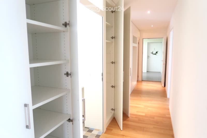 Empty hallway with wooden flooring, white walls, and an open closet with empty shelves on the left side.