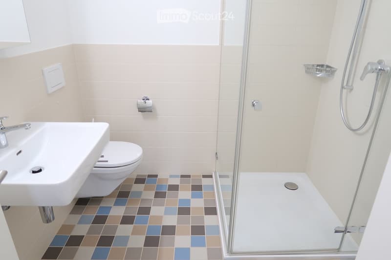Modern bathroom with a white sink, toilet, and glass-enclosed shower. The floor features a colorful tile pattern, and the walls are tiled in light beige.