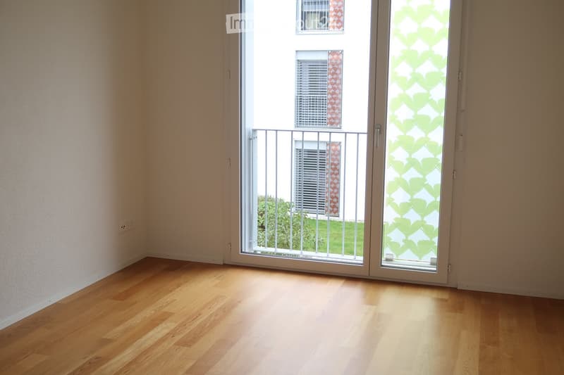 Empty room with wooden flooring, large sliding glass door with security bars, and a view of a building and greenery outside.
