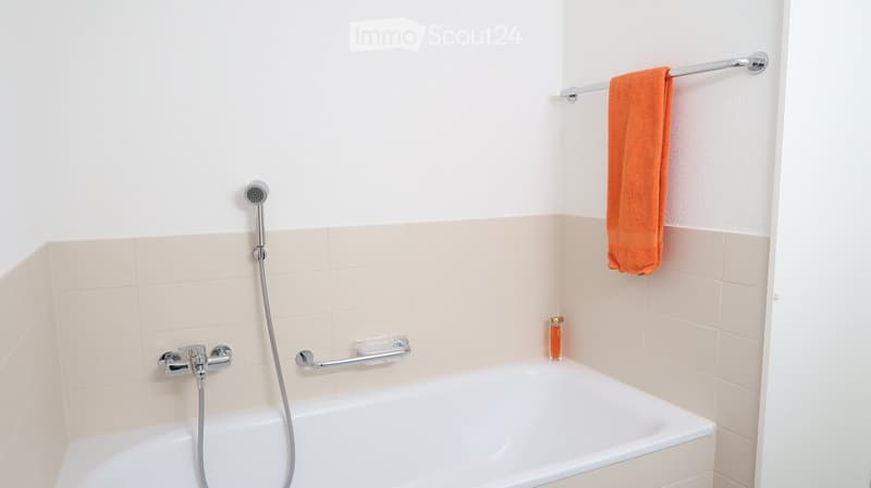 Modern bathroom with a bathtub, chrome faucet, handheld showerhead, towel rack with an orange towel, and a small bottle on the edge of the tub.