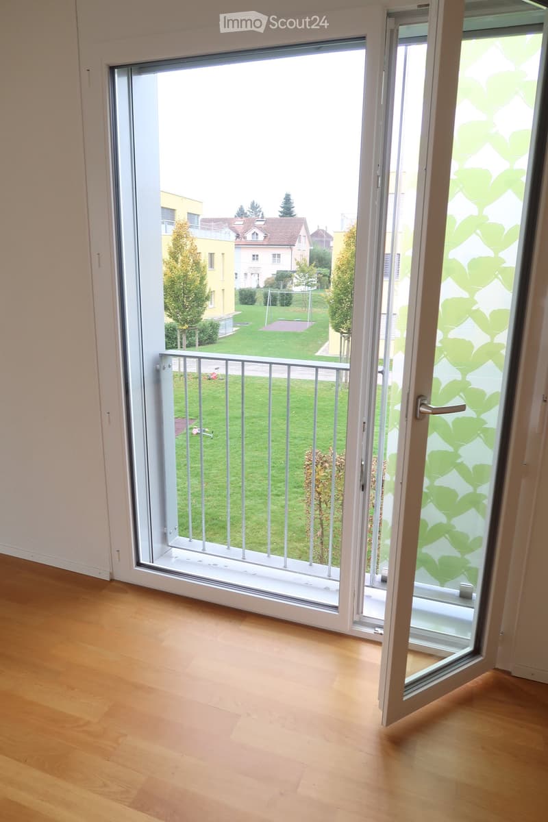 Empty room with wooden flooring and large sliding glass doors leading to a balcony overlooking a green lawn and neighboring houses.
