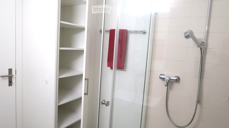Modern bathroom with white tiled walls, a glass-enclosed shower, a towel hanging on a rod, and an empty white cabinet with multiple shelves.
