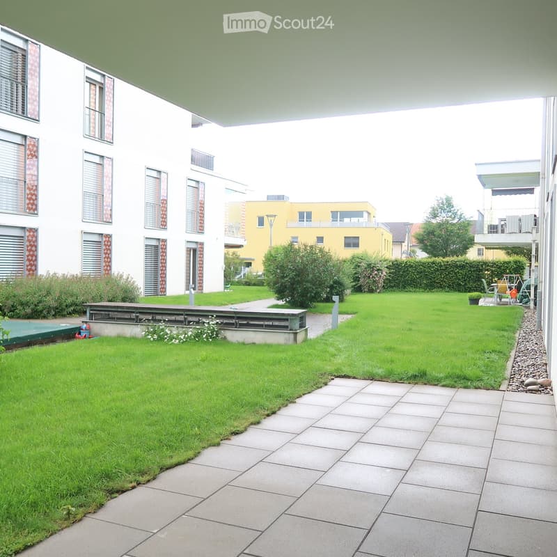 Well-maintained garden with green lawn, paved pathway, and a bench near the building entrance. The area is surrounded by modern residential buildings with large windows and balconies.