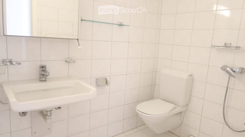 Modern bathroom with white tiles, a sink with a silver faucet, a toilet, a mirror, and glass shelves.