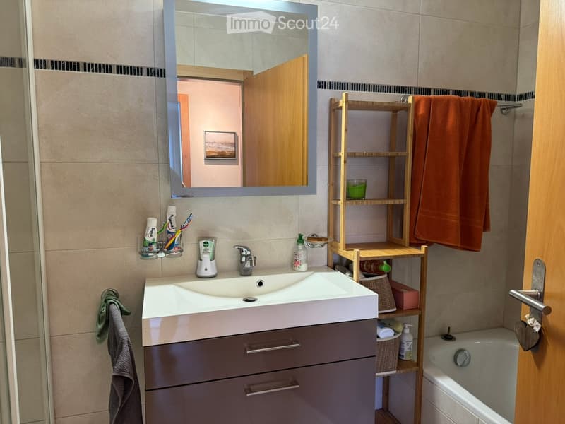 Modern bathroom with a white sink, mirror, wooden shelf, and bathtub. Includes a toothbrush holder, toothpaste, and towels.