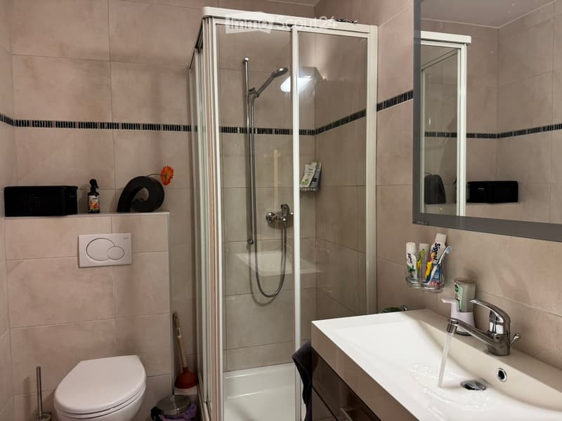 Modern bathroom with a white toilet, sink with running water, glass-enclosed shower, and a mirror above the sink. The walls are tiled in beige with black accents, and various toiletries and cleaning supplies are visible.