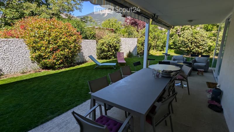 Outdoor patio with a long dining table, chairs, and a lounge area with a sofa and armchairs. The garden features a well-maintained lawn, lounge chairs, and a scenic mountain view.