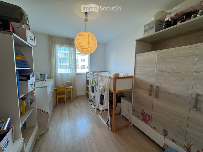 A well-lit bedroom with a bunk bed, a small desk with a yellow chair, shelves, and a large wooden wardrobe.