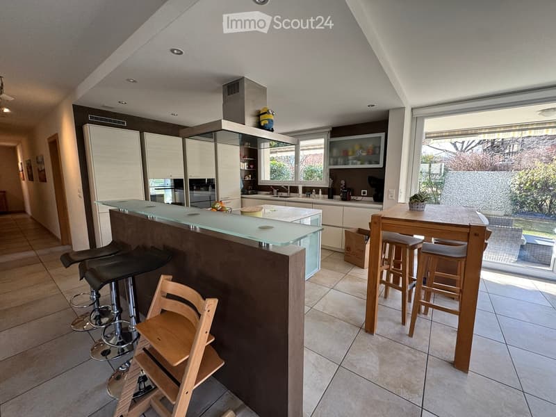 Modern kitchen with white cabinets, a central island with a glass countertop, bar stools, and a dining table with chairs. The space features large windows with blinds, a tiled floor, and a view of the garden.
