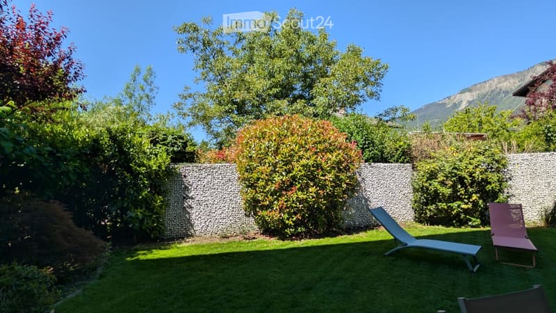 Well-maintained garden with green grass, stone wall, and lounge chairs, surrounded by lush trees and shrubs, with a mountain view in the background.
