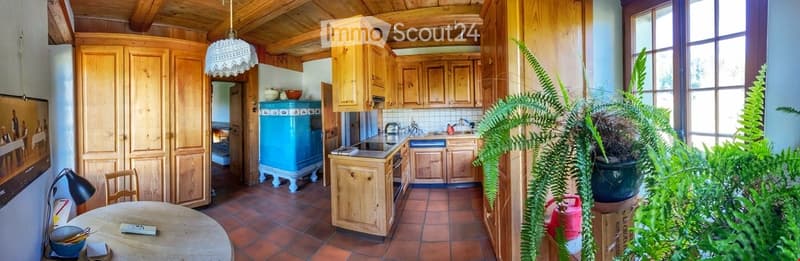 Bright kitchen with wooden cabinets, a round dining table, and a blue refrigerator. The space features a sink, oven, and a hanging lamp, with large windows letting in natural light.