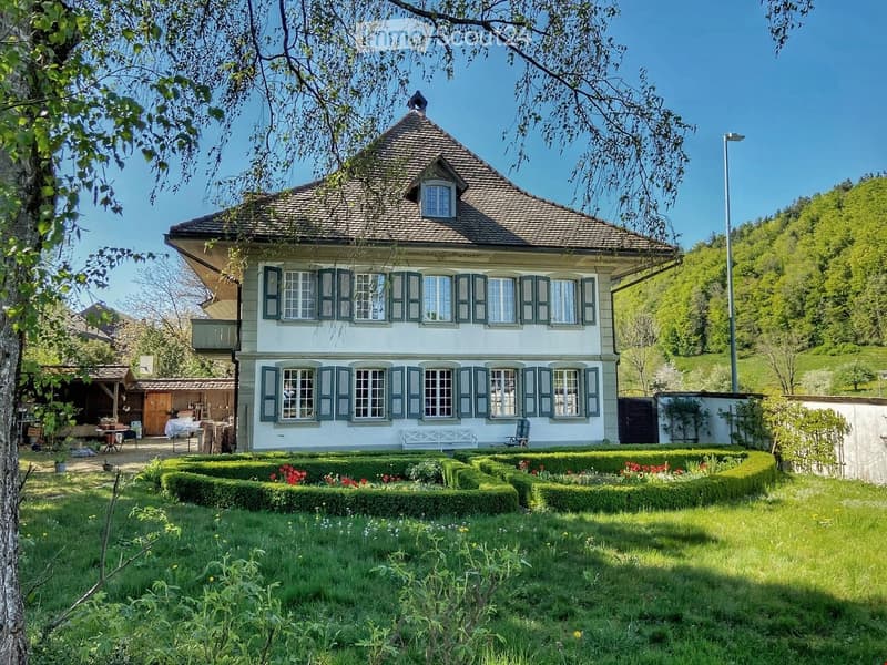 A two-story house with a traditional Swiss architectural style, featuring a pitched roof, multiple windows with shutters, and a well-maintained garden with neatly trimmed hedges and flower beds.