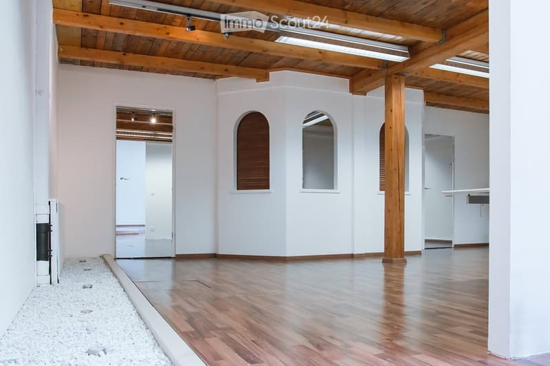Empty room with white walls, wooden ceiling beams, and hardwood flooring. Features arched windows and a small seating area with a table and chairs.