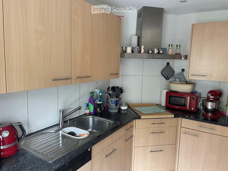Modern kitchen with light wooden cabinets, stainless steel sink, red kettle, microwave, mixer, and various kitchen utensils on the counter.