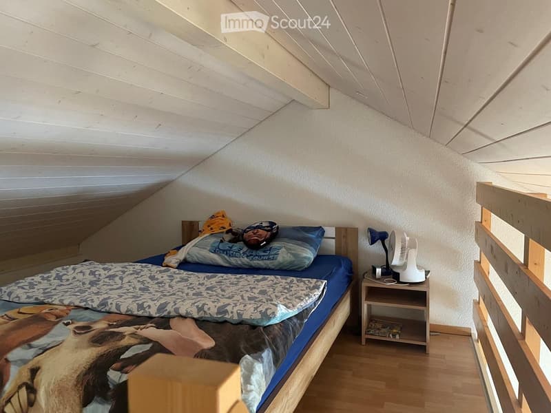 Cozy attic bedroom with wooden slanted ceilings, a single bed with blue bedding, a nightstand with a fan and lamp, and a small wooden railing.