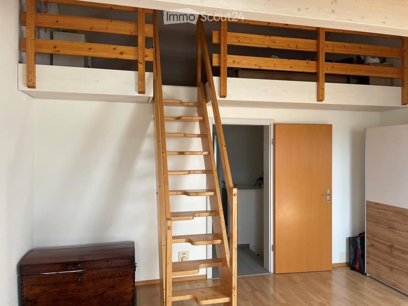 A compact room with a wooden staircase leading to a loft area, featuring a wooden chest and a closed door leading to another space.