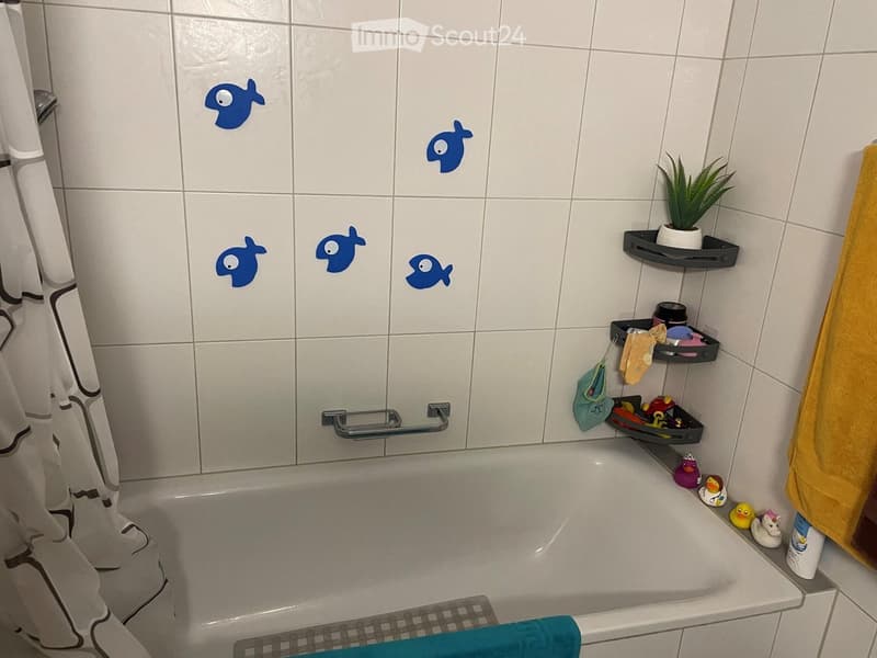 Bathtub with a colorful sticker design on the tiles, a towel hanging on the side, shelves holding various items including a plant, toys, and a towel.