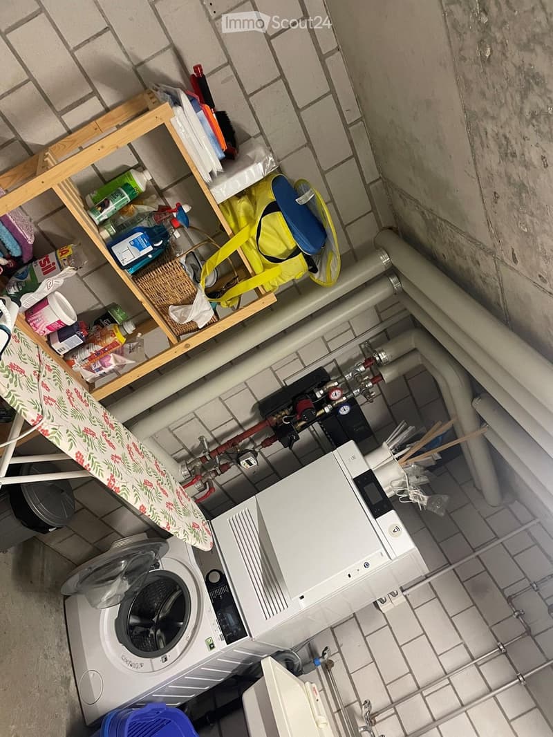 Laundry area with a washing machine, ironing board, cleaning supplies, and visible pipes.