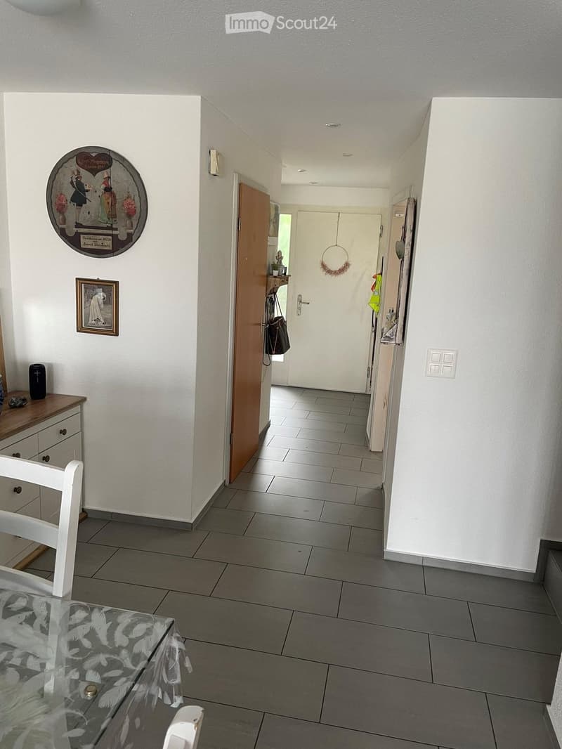 A clean, modern hallway with tiled flooring, white walls, and several doors leading to other rooms. There are decorative items on the walls, a small table with a speaker, and a coat rack with a hanging bag.