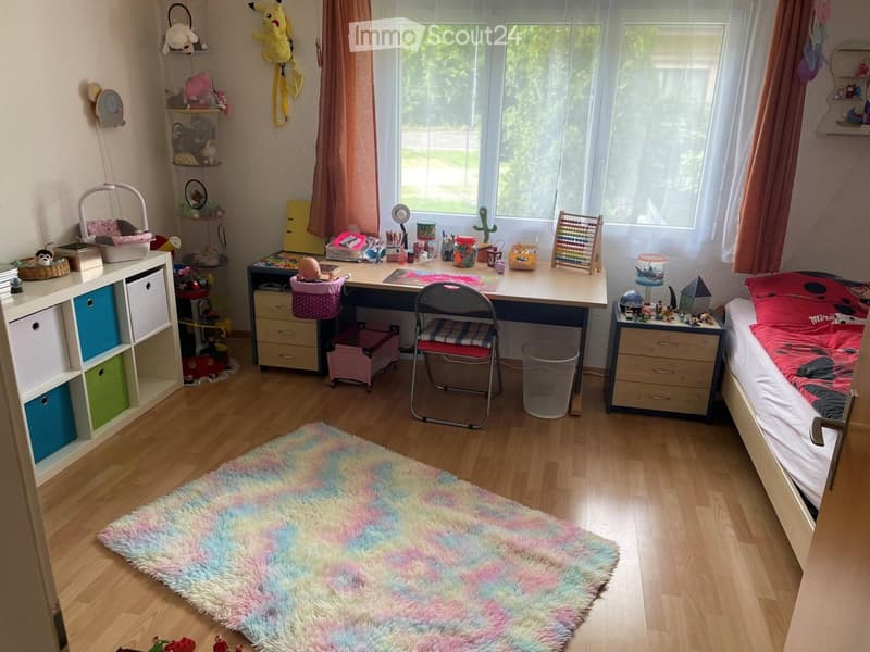 A well-lit children's bedroom with a bed, desk, and various toys. The room features a colorful shaggy rug, a storage unit, and a window with curtains.