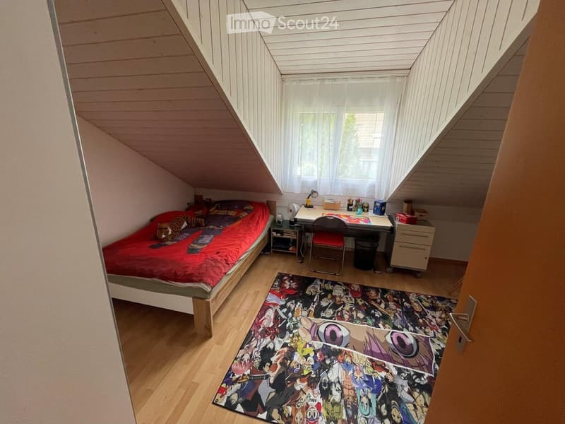 Cozy attic bedroom with slanted wooden ceiling, bed with red bedding, desk with chair, and colorful anime-themed rug.