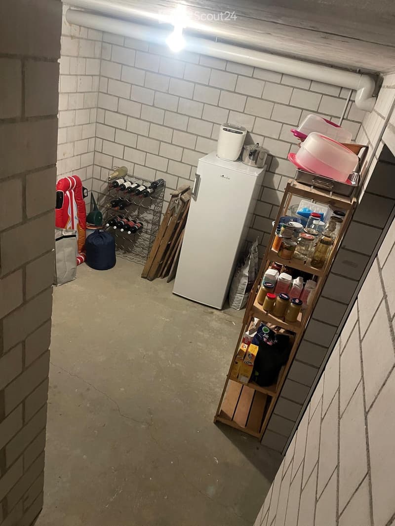 A small, well-lit basement storage area with a refrigerator, wine rack, wooden shelf, and various storage bins.