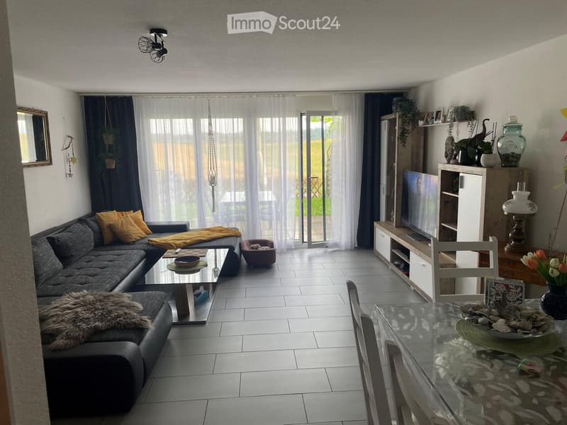 Modern living room with a grey sectional sofa, coffee table, glass dining table, TV stand, and decorative items including a vase with flowers and a lamp.